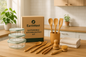 Create a bright, modern kitchen scene showcasing the EarthNest Sustainable Kitchen Essentials Kit. Arrange all items — reusable glass storage containers, bamboo utensils, organic cotton dish towels, and biodegradable cleaning brushes — neatly on a wooden countertop. Include soft natural lighting, green plants, and neutral kitchen decor to emphasize eco-friendliness. The scene should feel warm, inviting, and lifestyle-oriented, highlighting the complete set of products together.